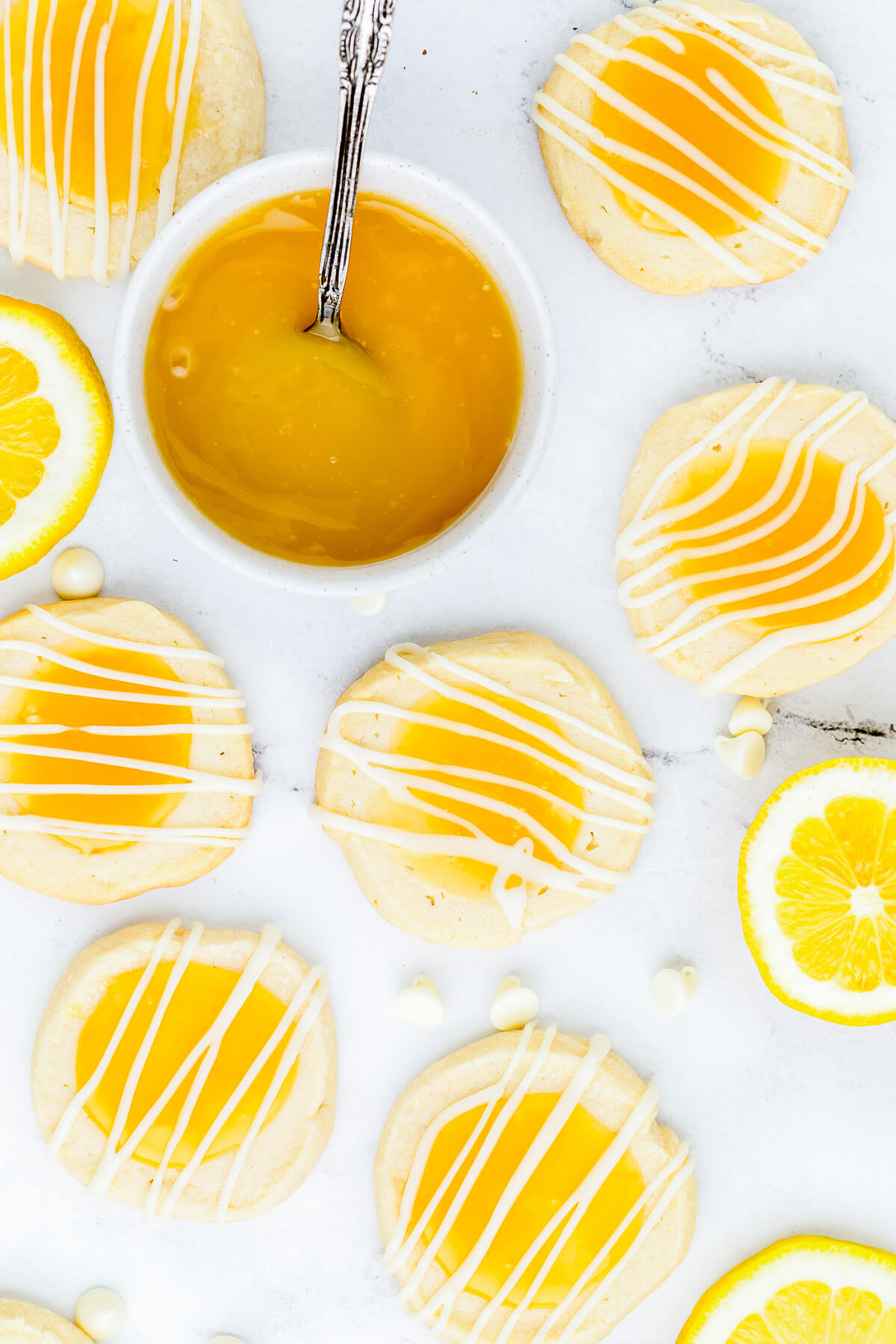 A group of Lemon curd thumbprint cookies topped with a white chocolate drizzle with a small bowl of lemon curd on white marble. 