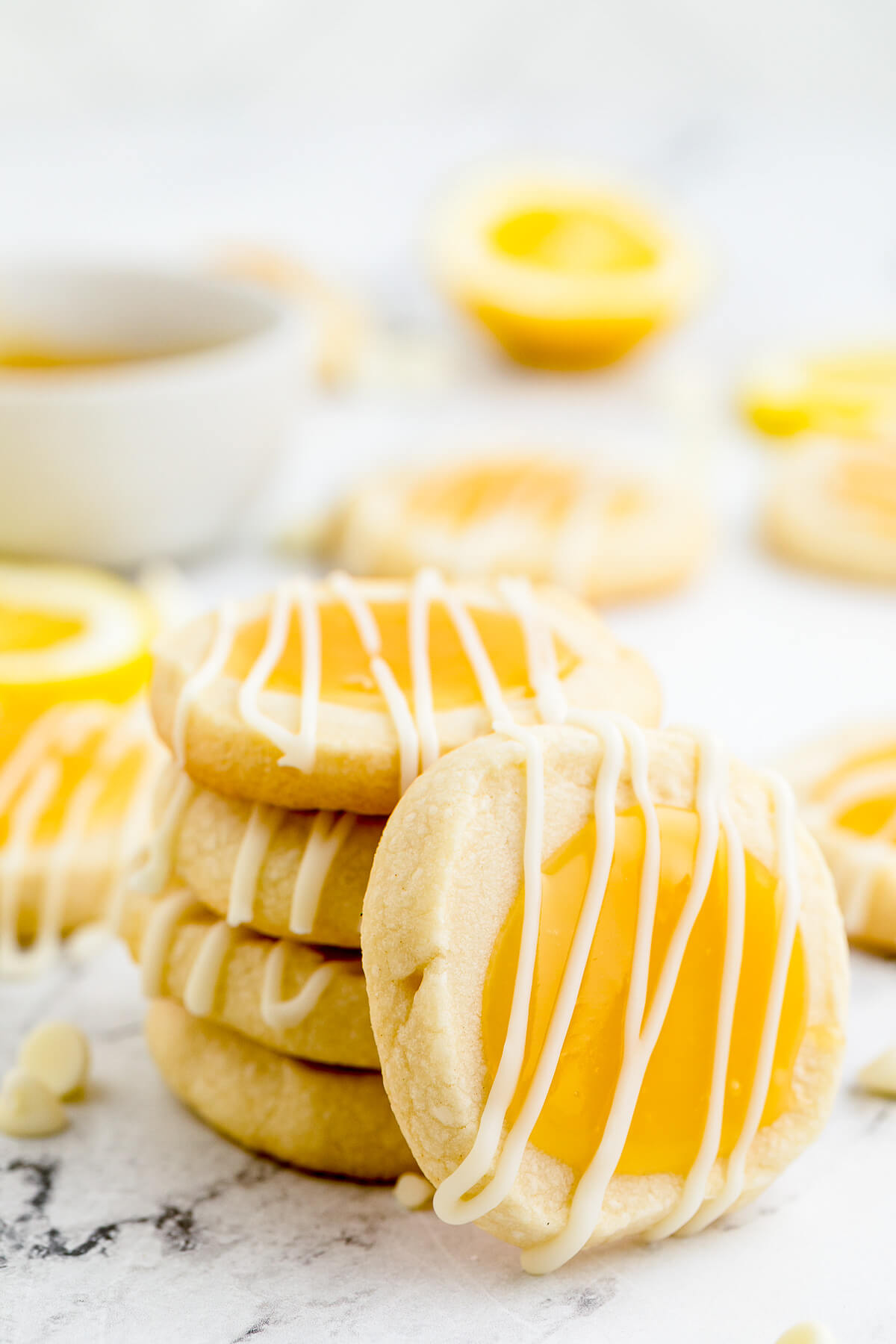 Four stacked Lemon curd thumbprint cookies topped with a white chocolate drizzle with another leaning against the stack.