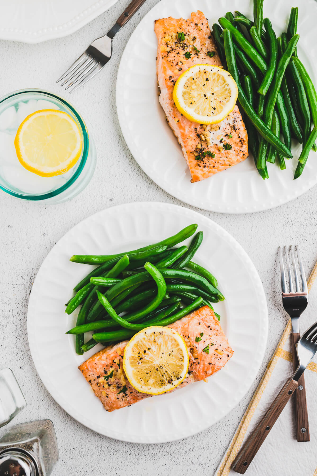 A table scene with two dinner plates of baked lemon pepper salmon with green beans surrounded by lemons, forks, and napkins.