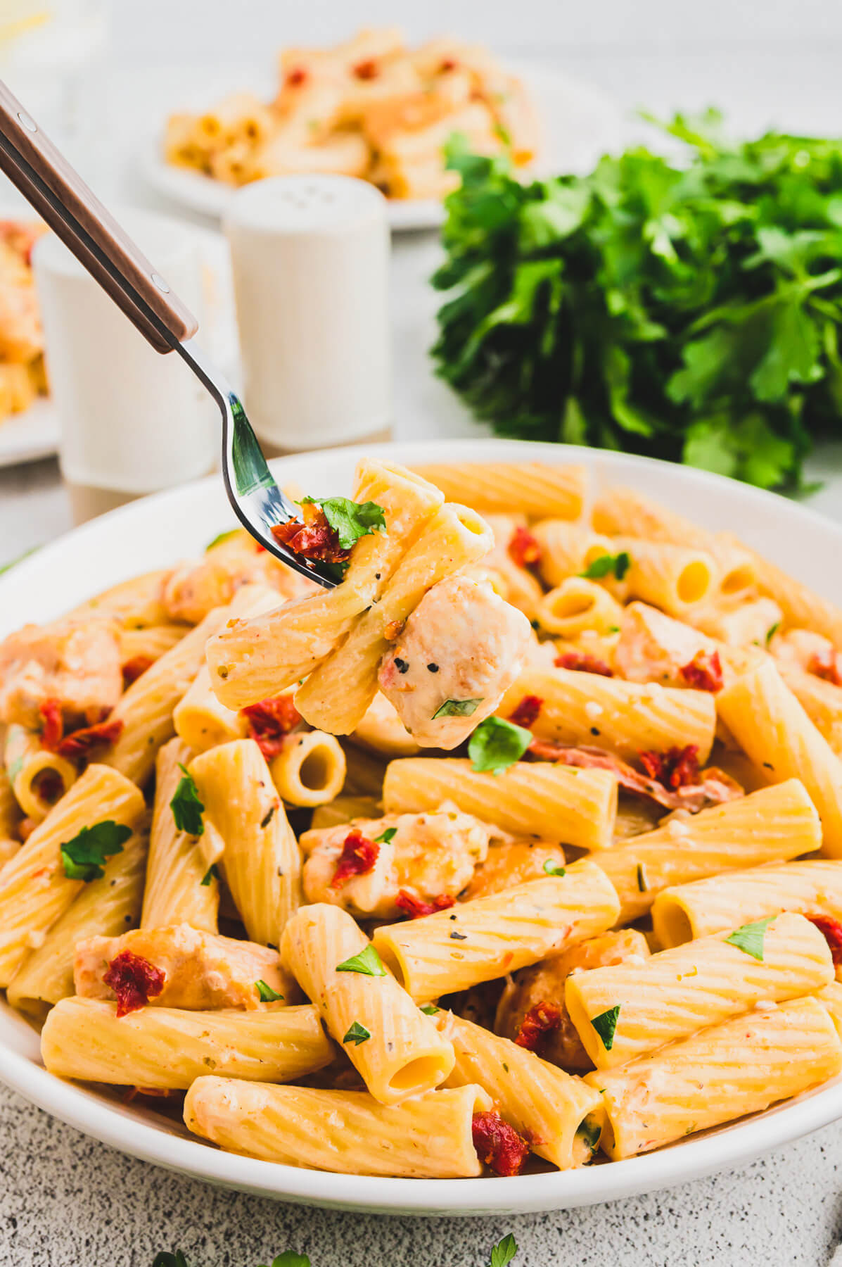 A fork above a white bowl of Marry Me Chicken Pasta featuring rigatoni pasta, chicken, sun dried tomatoes, and herbs in a creamy pasta sauce.