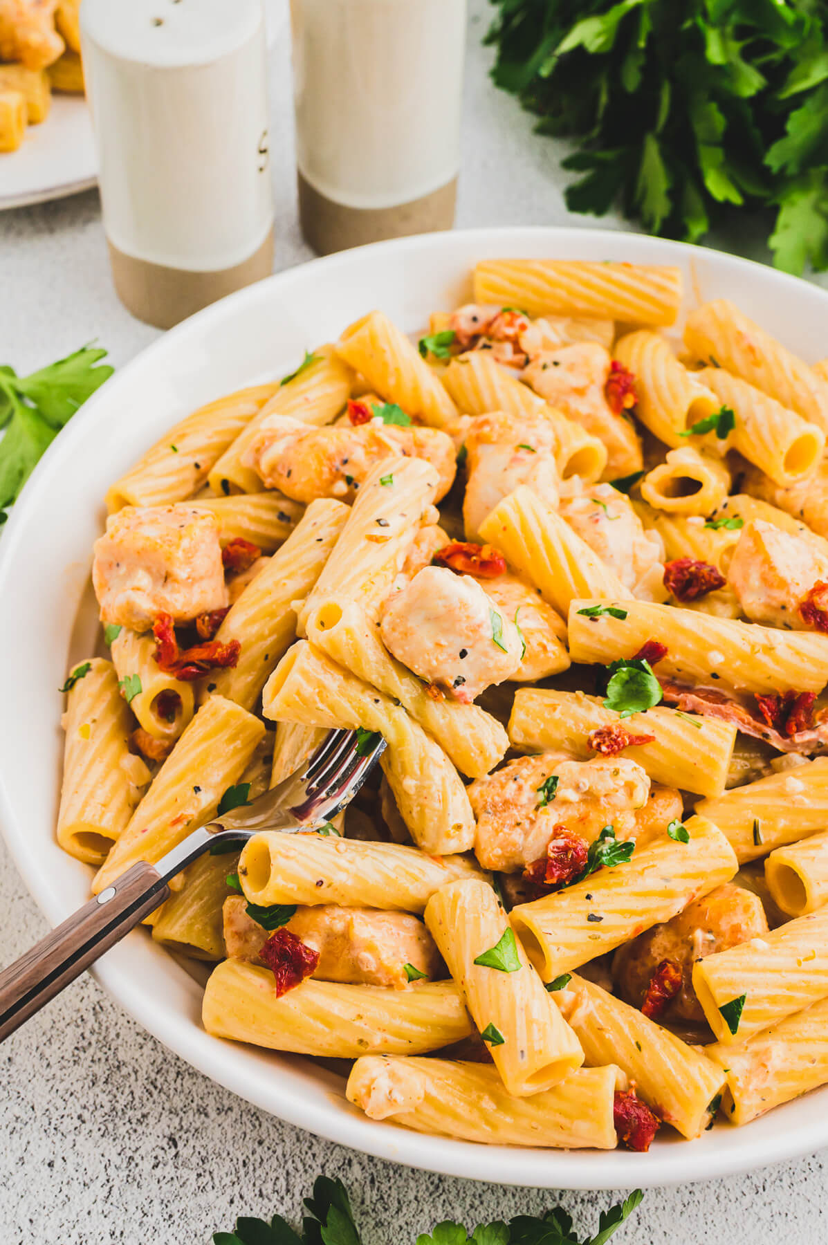 A fork in a white bowl of Marry Me Chicken Pasta featuring rigatoni pasta, chicken, sun dried tomatoes, and herbs in a creamy pasta sauce.