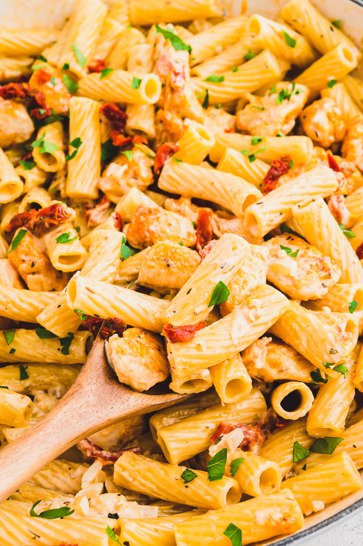 A wooden spoon full of rigatoni pasta, chicken, sun dried tomatoes, and herbs in a creamy pasta sauce.