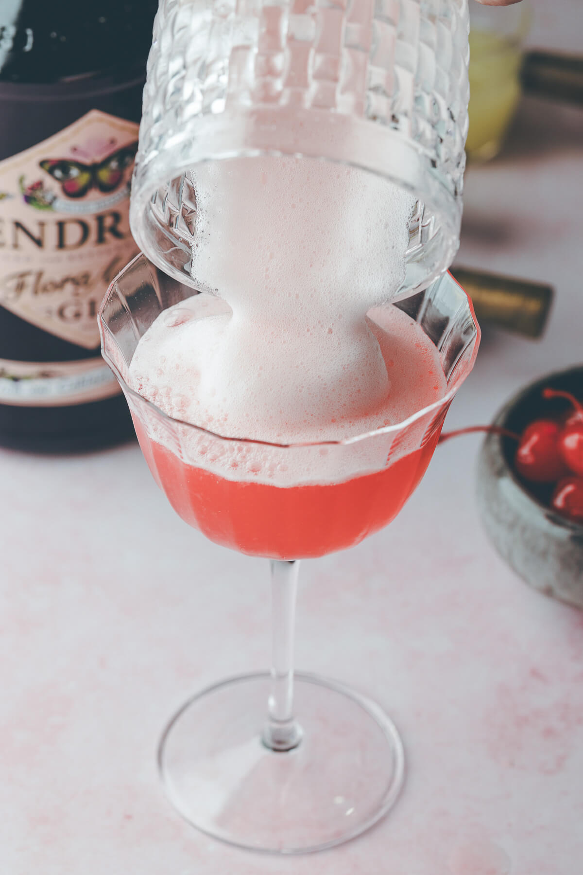 Adding egg white foam to a pink lady cocktail in a coupe glass.