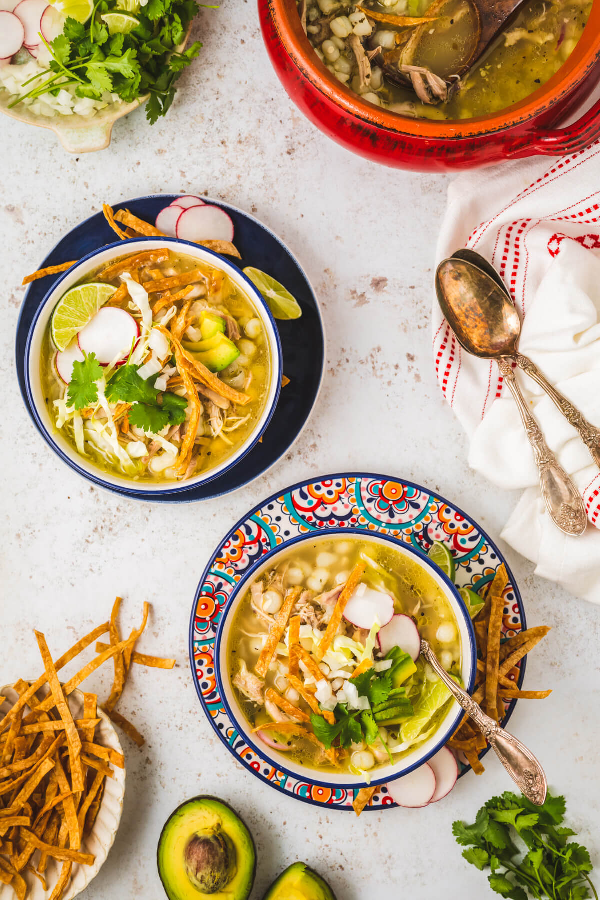 A table scene featuring two colourful soup bowls containing Pozole blanco featuring white hominy, cilantro, radishes, cabbage, lime wedge, and strips of fried tortilla.