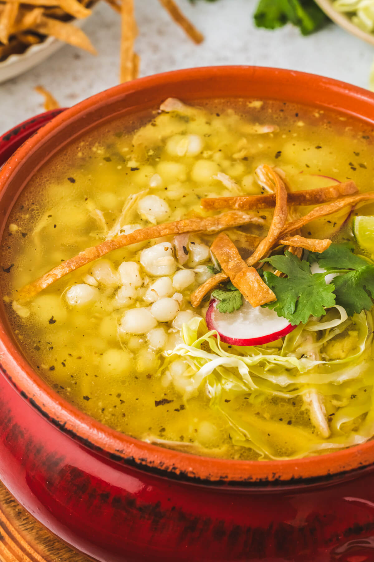 A bright terra cotta pot filled with Pozole blanco featuring white hominy, cilantro, radishes, cabbage, lime wedge, and strips of fried tortilla.