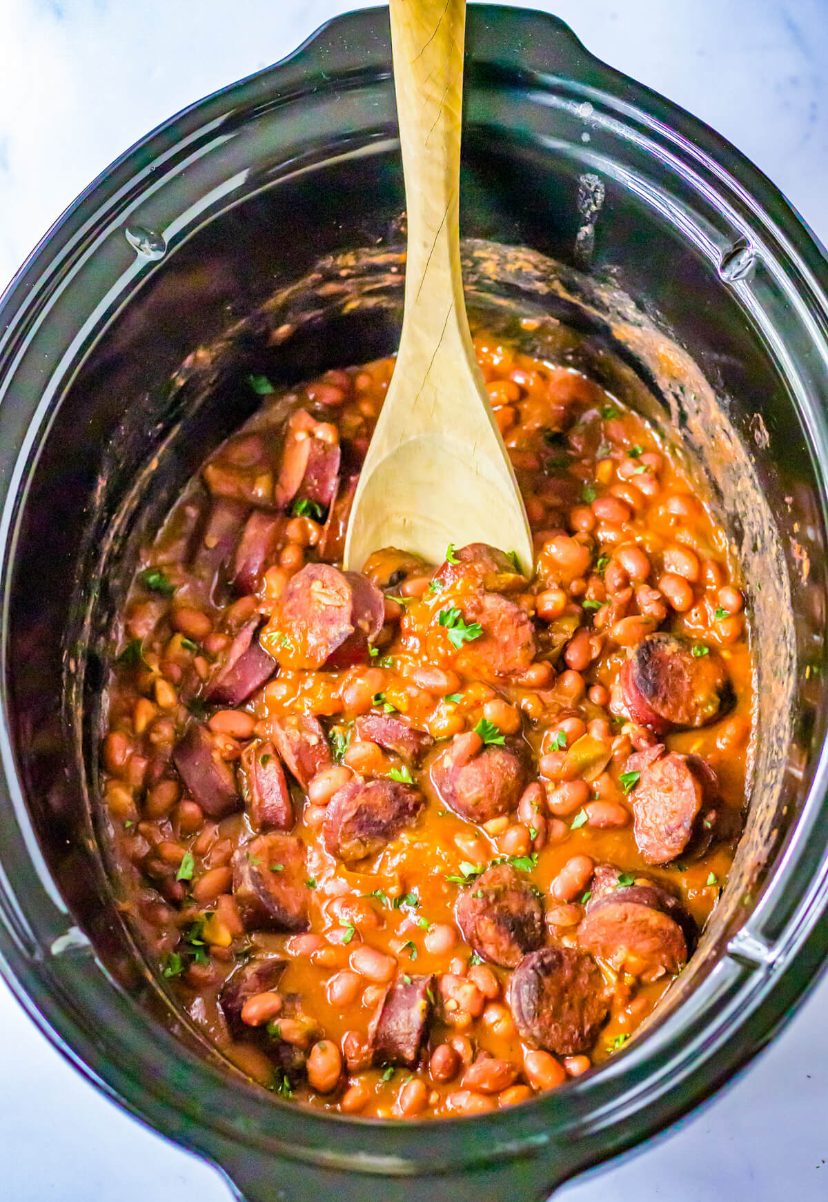 A crock pot insert containing a wooden spoon sticking into the cooked red beans and rice.