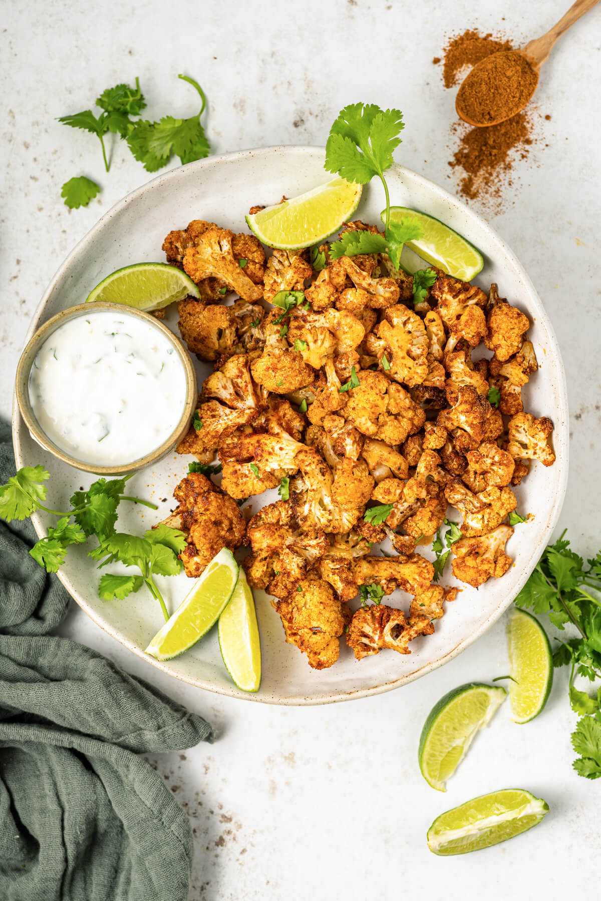 A plate of vibrant curried air fryer cauliflower beside a tiny bowl of creamy dip, cilantro, and lime wedges.