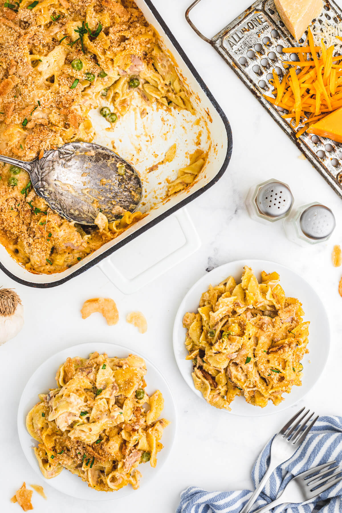 A white enamel baking pan half full of golden baked, crumb topped Tuna Noodle Casserole beside two full white plates.
