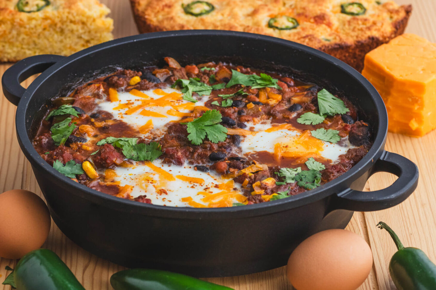 A cast iron skillet full of baked chili and eggs surrounded by cornbread, cheddar cheese, eggs, and jalapenos.