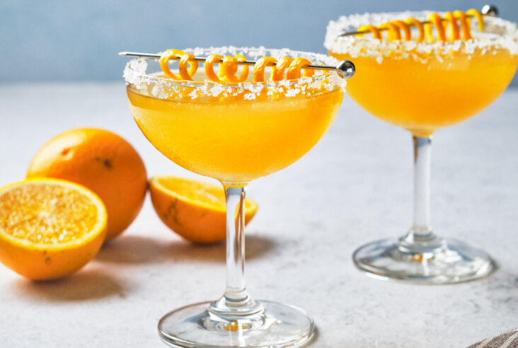 Two coupe glasses rimmed with salt containing orange Italian Margarita cocktail garnished with coiled orange peels on metal cocktail picks.