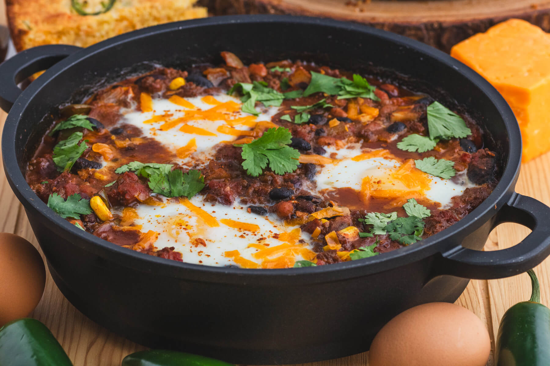 A cast iron skillet full of baked chili and eggs surrounded by cornbread, cheddar cheese, eggs, and jalapenos.
