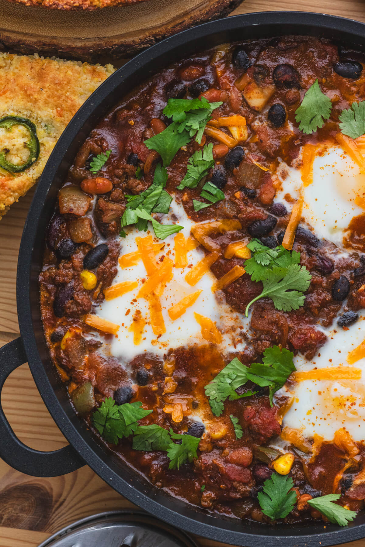 A cast iron skillet full of baked chili and eggs beside jalapeno cheddar cornbread.