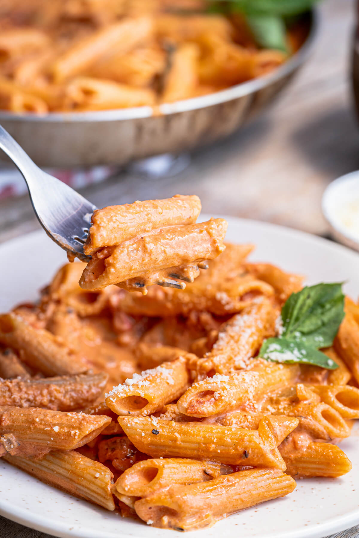 A fork filled with penne pasta in Rosé sauce garnished with grated Parmesan and fresh basil leaves.