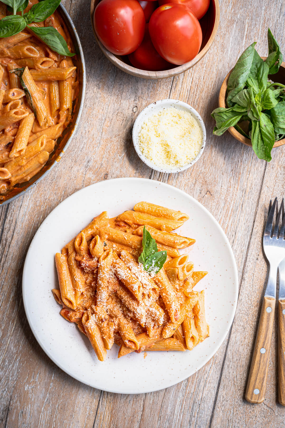 A white plate with a serving of cooked penne pasta in Rosé sauce garnished with grated Parmesan and fresh basil leaves.