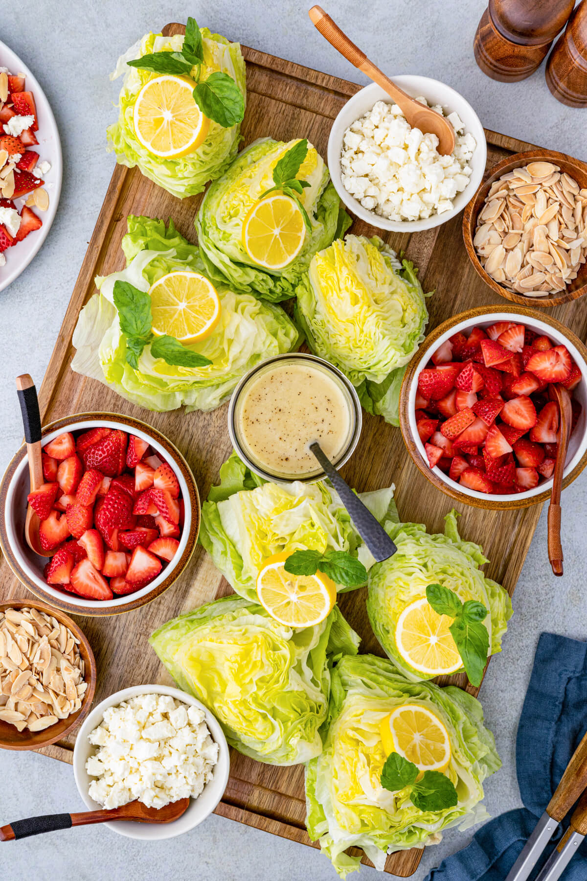 Iceberg lettuce wedges and lemon vinaigrette arranged on a wooden serving board with bowls of strawberries, feta, and almonds.