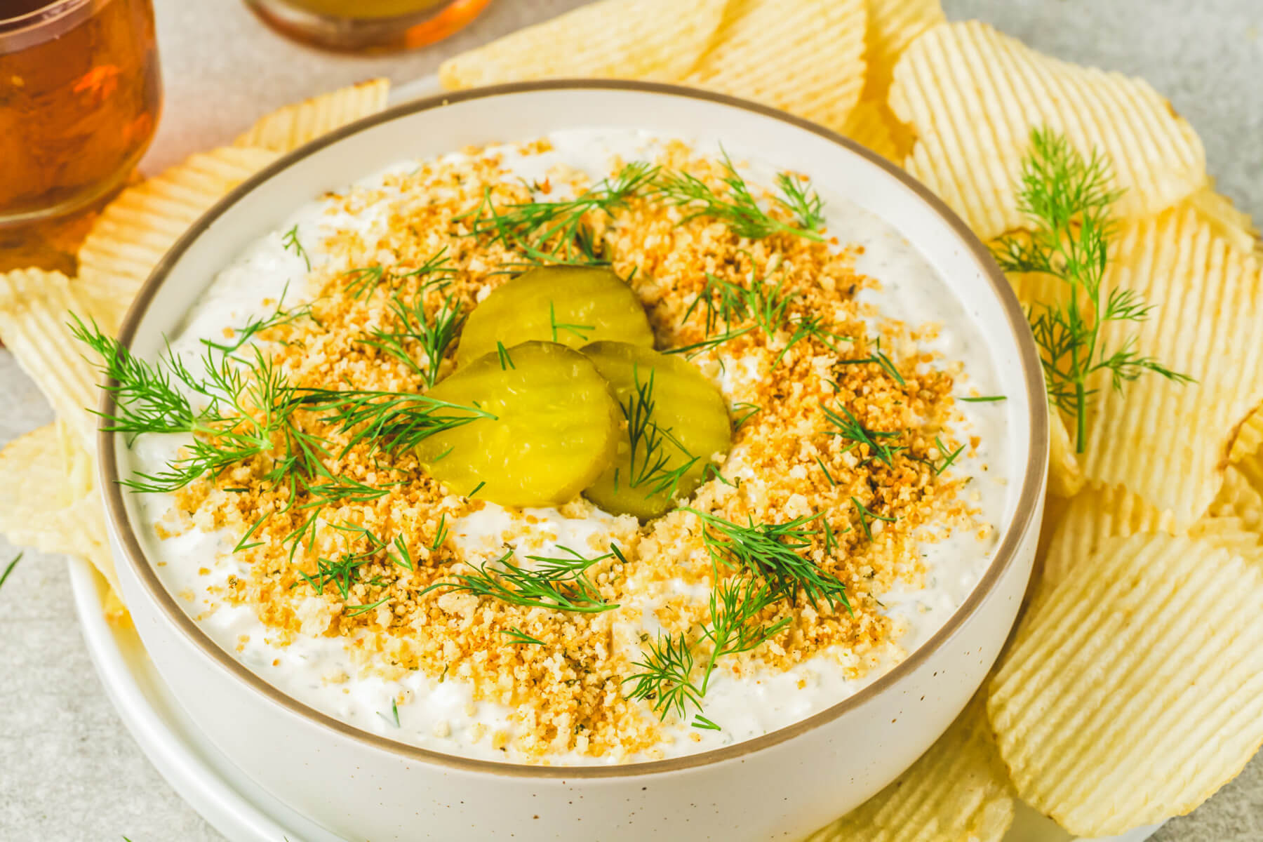 Ridged potato chips surround a bowl of white dill pickle dip topped with golden panko crumbs, fresh dill, and sliced dill pickles.