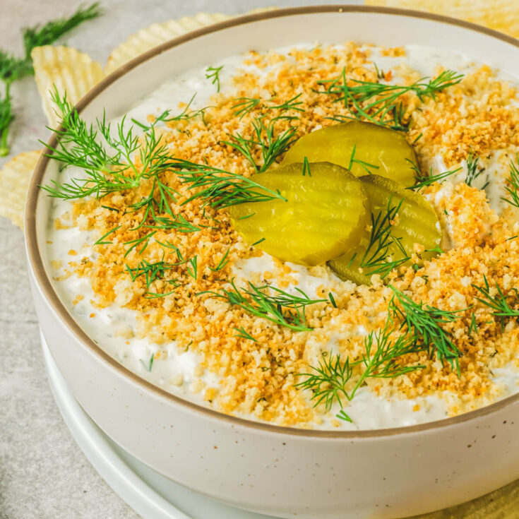Ridged potato chips surround a bowl of white dill pickle dip topped with golden panko crumbs, fresh dill, and sliced dill pickles.