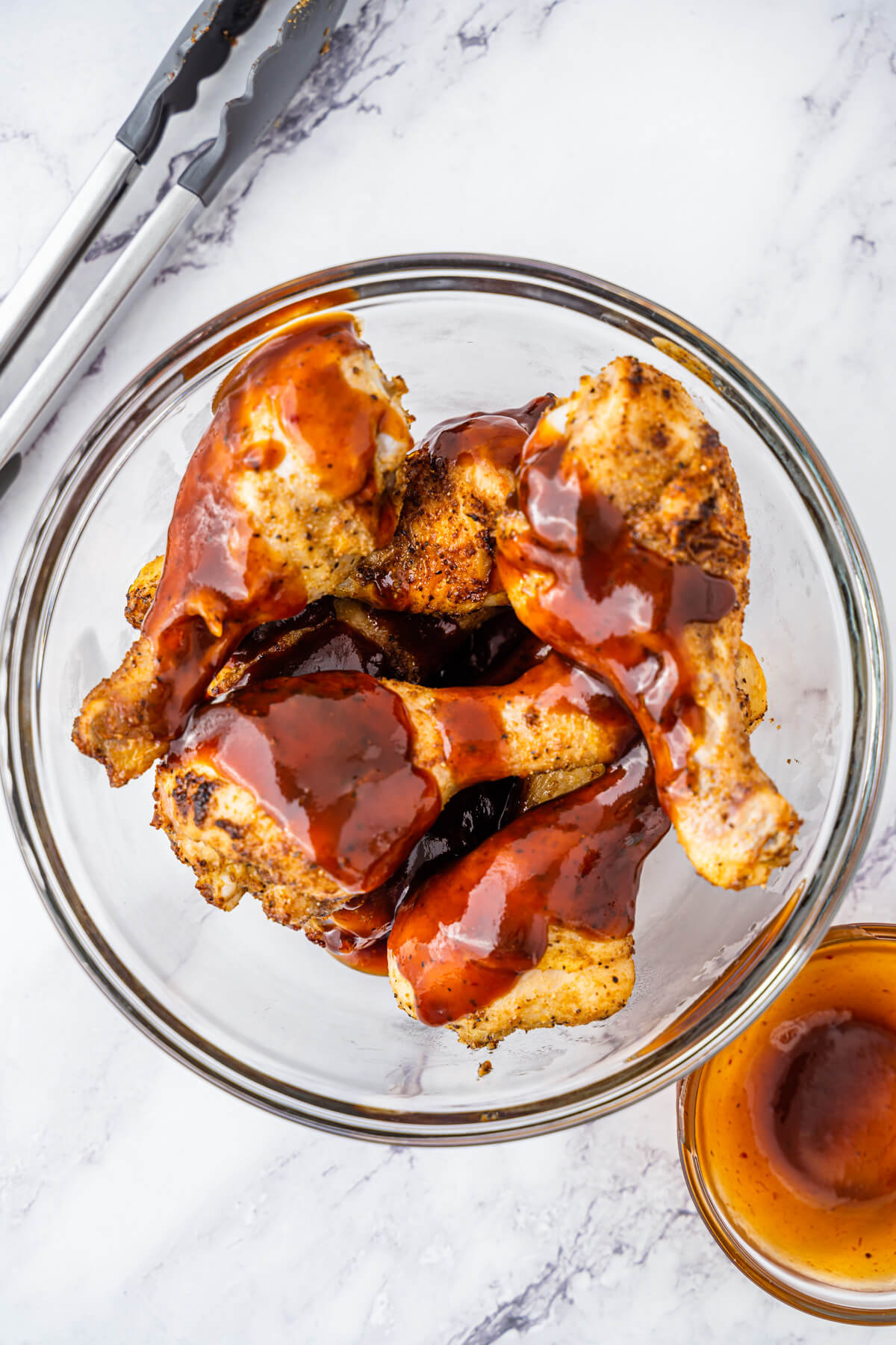 A glass bowl containing  chicken legs coated in barbecue sauce.