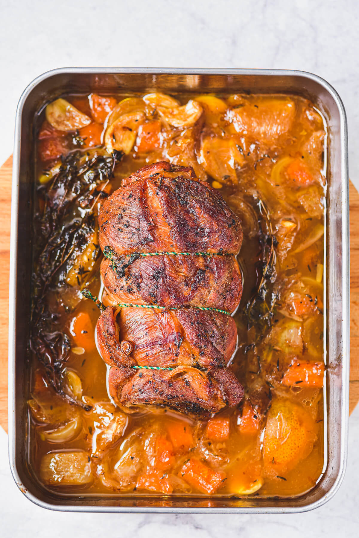 A fully cooked rolled lamb breast roast in a roaster surrounded by beef stock, vegetables, and fresh thyme leaves.