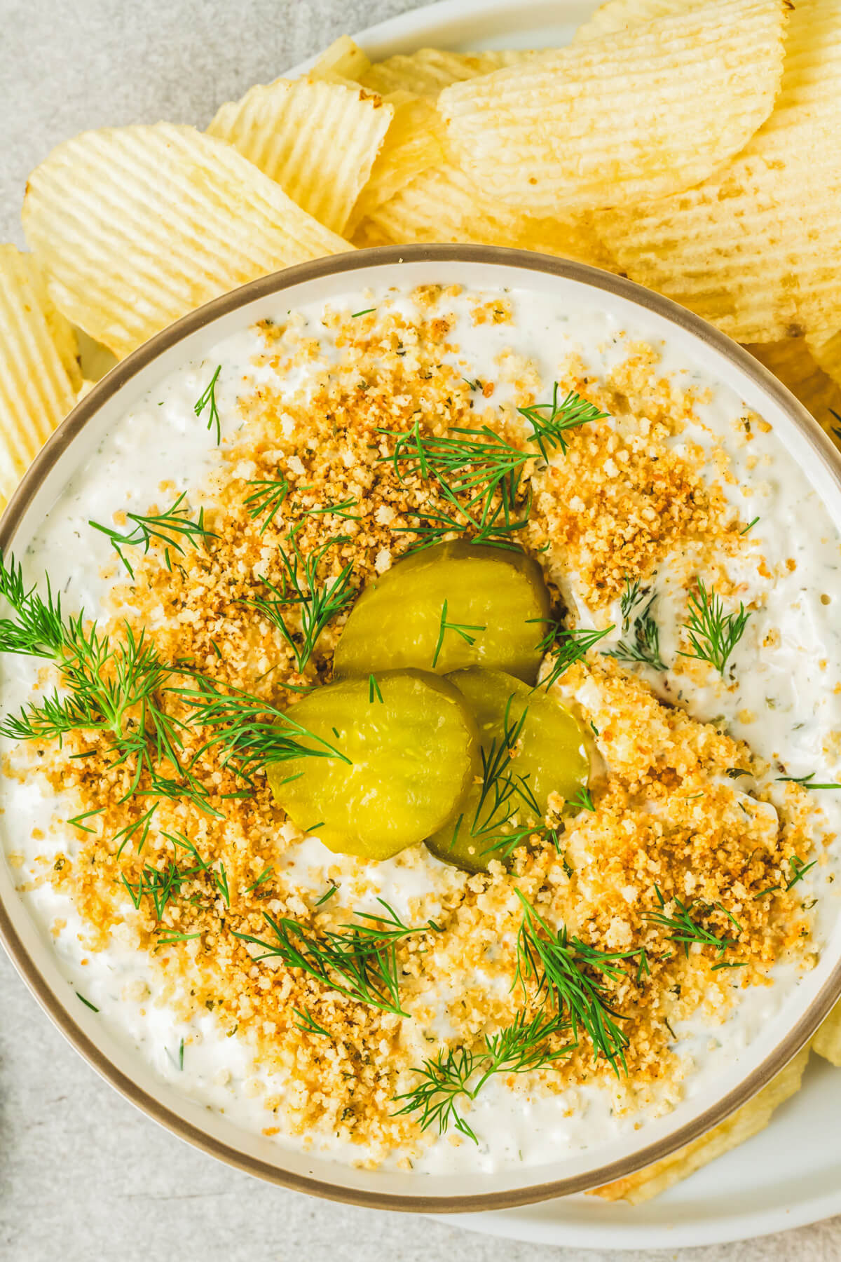 Ridged potato chips surround a bowl of white dill pickle dip topped with golden panko crumbs, fresh dill, and sliced dill pickles. 