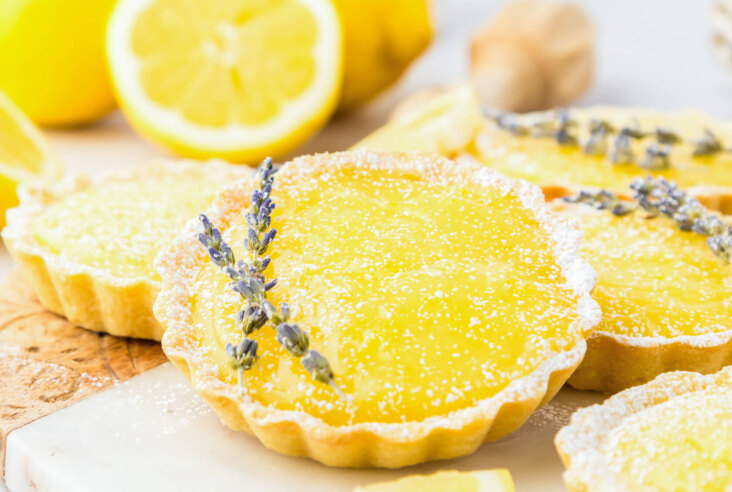 A bright yellow lemon curd filled tart garnished with two sprigs of lavender beside a cut lemon and more lemon tarts.