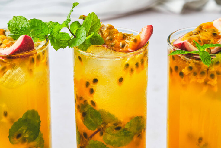 Three bold Passion Fruit Mojitos in tall highball glasses garnished with sprigs of mint, purple passion fruit wedges, and pulp.
