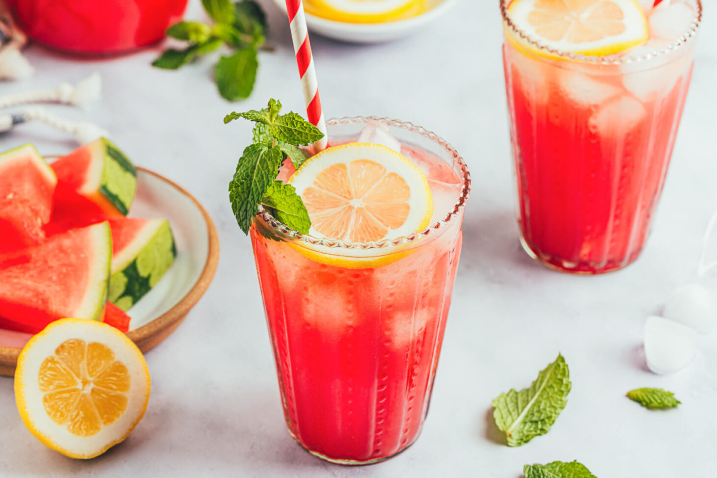 Two glasses of vibrant pink Watermelon Mint Lemonade garnished with fresh mint and lemon slices beside a plate of sliced watermelon.