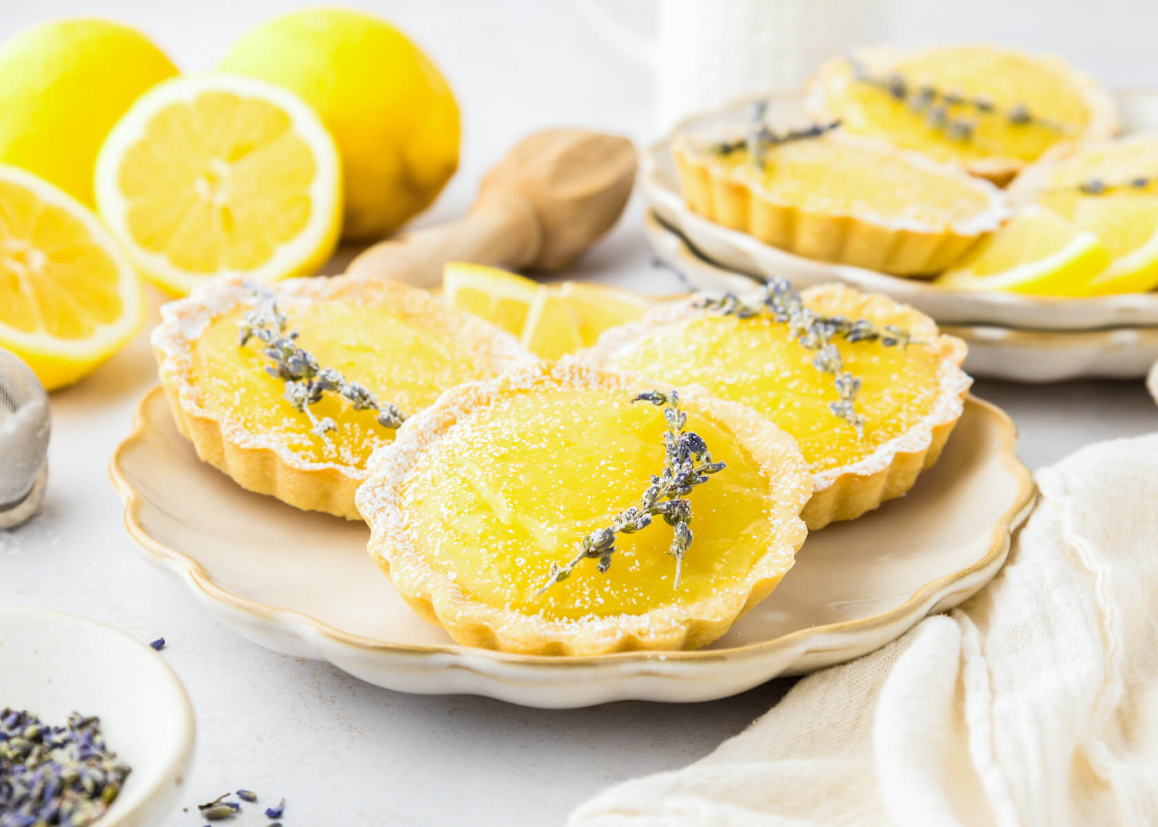Three bright yellow lemon curd filled tarts garnished with two sprigs of lavender on a plate beside a cut lemon and more lemon tarts.