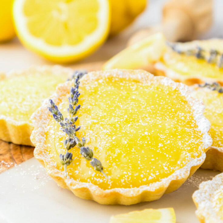 A bright yellow lemon curd filled tart garnished with two sprigs of lavender beside a cut lemon and more lemon tarts.