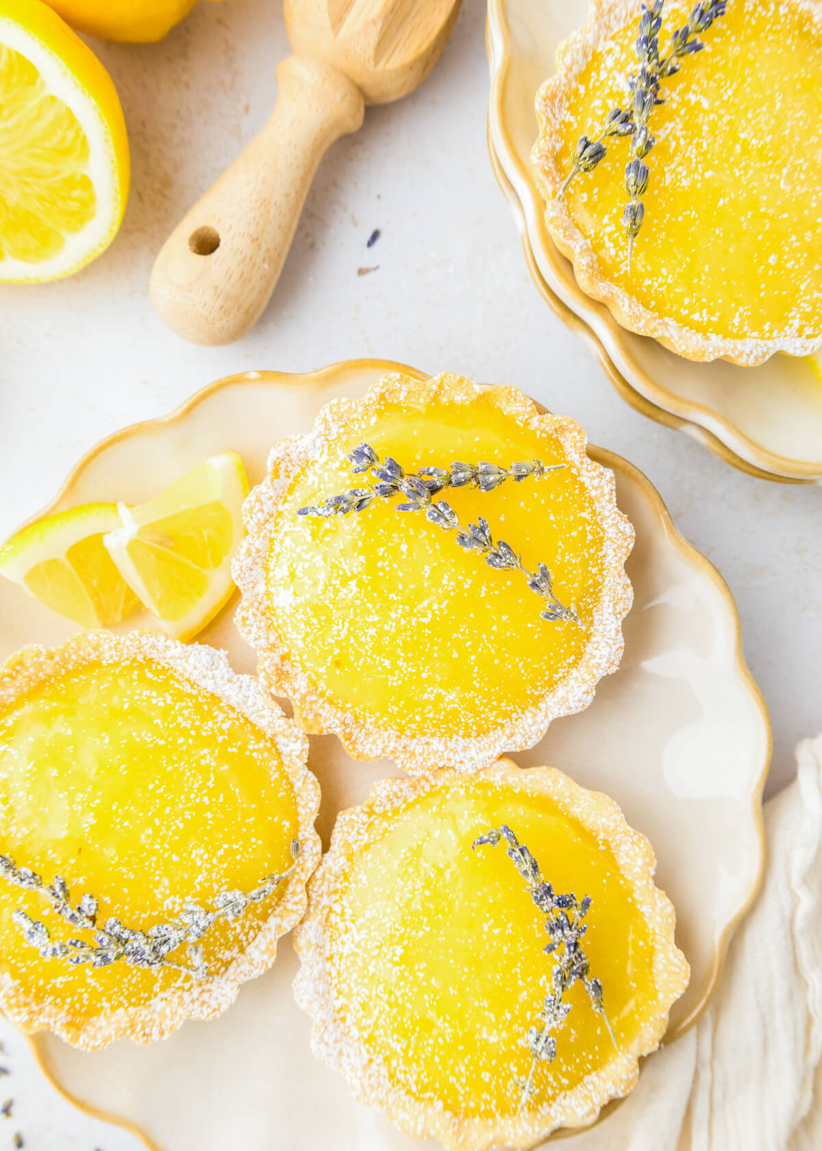 Three bright yellow lemon curd filled tarts each garnished with two sprigs of lavender beside a cut lemon and wooden lemon juicer.