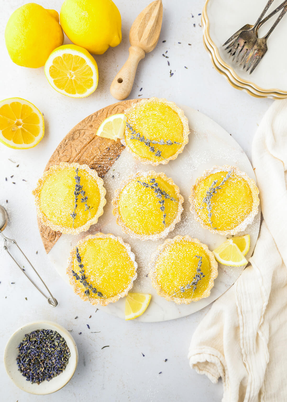 Six bright yellow lemon curd filled tarts garnished with two sprigs of lavender on a plate beside a cut lemons a wooden citrus reamer and a small bowl of lavender.