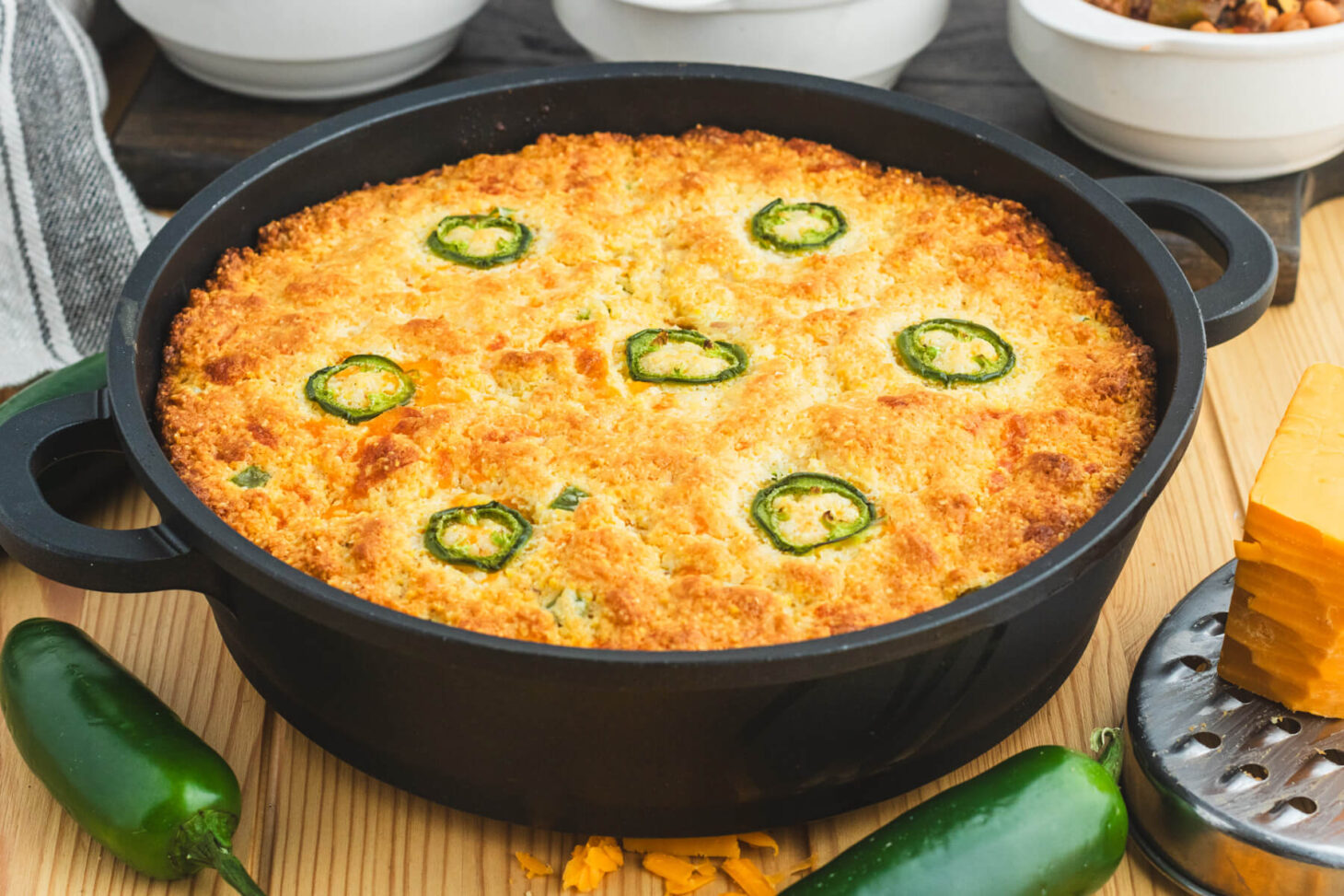 A cast iron skillet filled with golden baked cornbread topped with sliced jalapenos and surrounded by bowls of chili, grated cheese, and whole jalapenos.