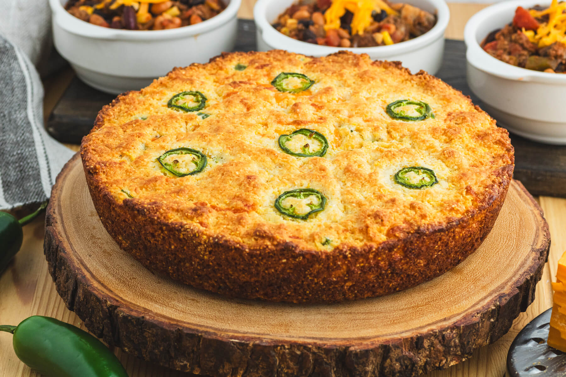 Golden baked cornbread topped with sliced jalapenos and surrounded by bowls of chili, grated cheese, and whole jalapenos.