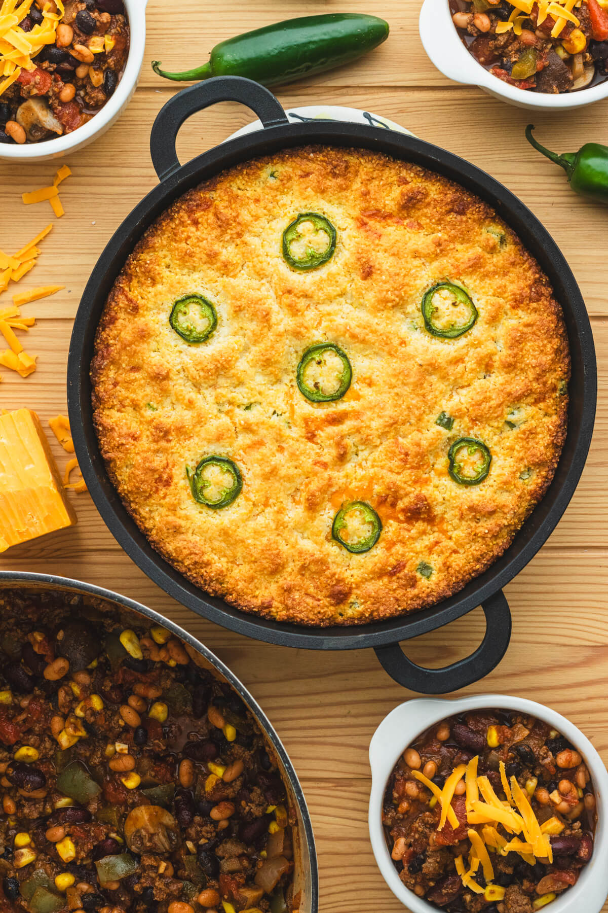 A cast iron skillet filled with golden baked cornbread topped with sliced jalapenos and surrounded by bowls of chili, grated cheese, and whole jalapenos.