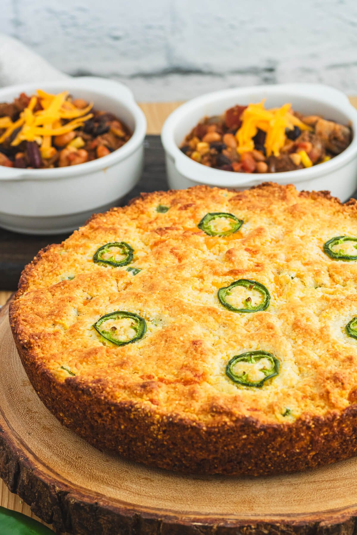 Golden baked cornbread topped with sliced jalapenos and surrounded by bowls of chili, grated cheese, and whole jalapenos.