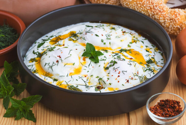 A black bowl of Turkish Eggs, or Çilir, featuring garlic yogurt, poached eggs, vibrant fresh herbs, and bright orange chili oil.