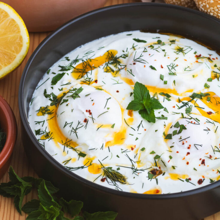 A black bowl of Turkish Eggs, or Çilir, featuring garlic yogurt, poached eggs, vibrant fresh herbs, and bright orange chili oil.