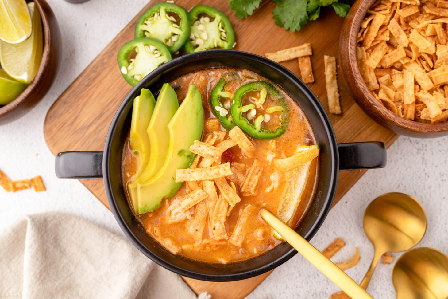 A black soup bowl full of creamy Chicken Enchilada Soup garnished with sliced avocados, tortilla strips, cilantro, lime, and jalapenos.