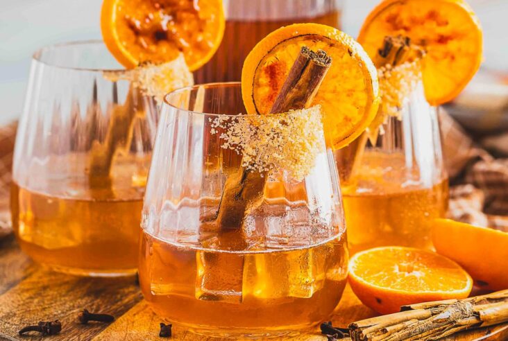 Three bourbon old fashioned cocktails in a rocks glasses garnished with orange slices and cinnamon sticks on a wooden board.