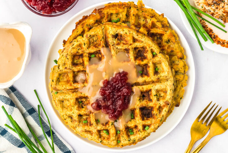 A white plate containing two crispy golden Stuffing Waffles topped with gravy and cranberry sauce.