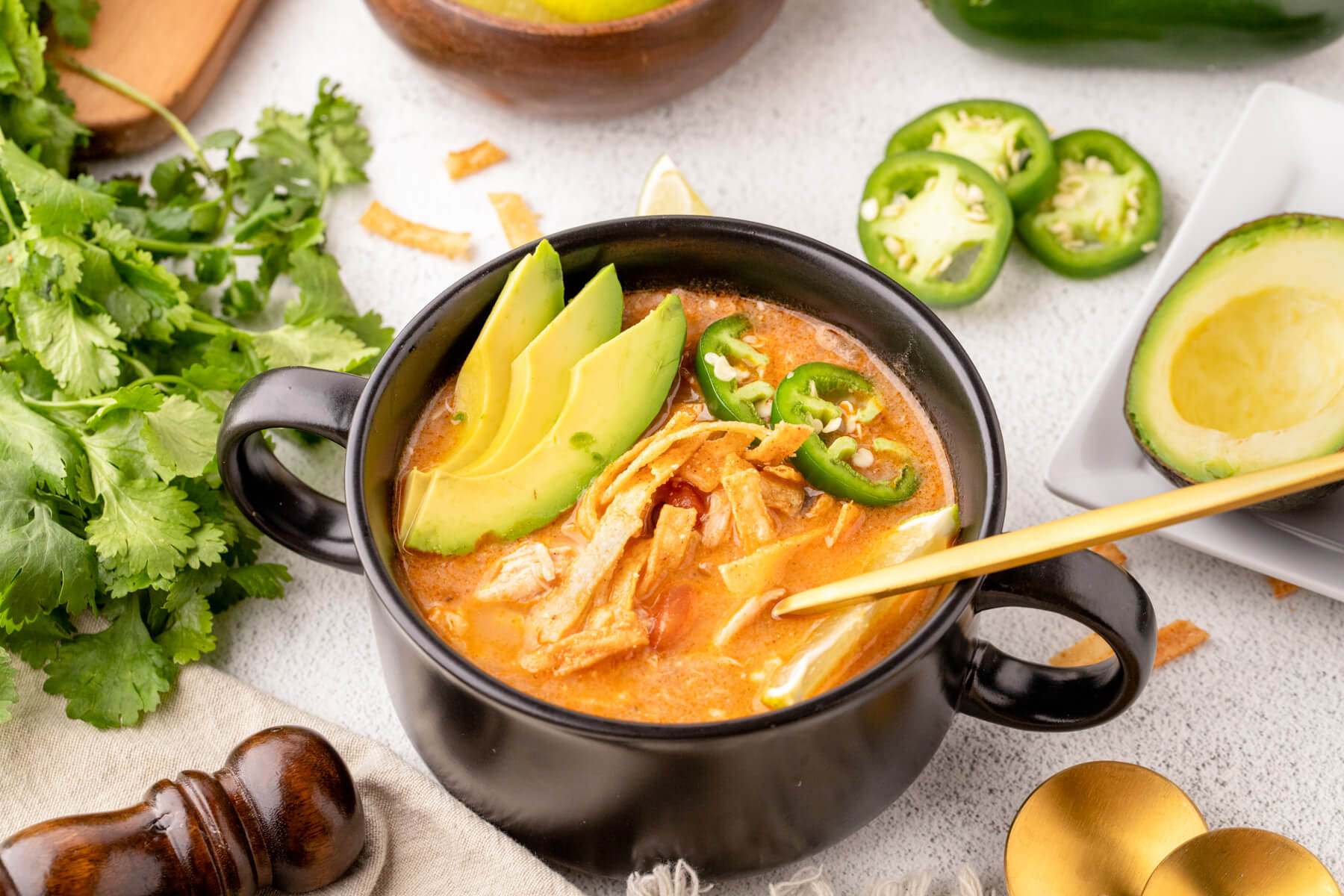 A black soup bowl full of creamy Chicken Enchilada Soup garnished with sliced avocados, tortilla strips, cilantro, lime, and jalapenos.