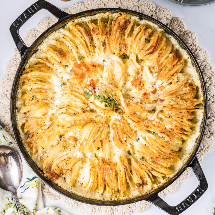 A large Staub baking dish containing golden baked cheesy potatoes au gratin and fresh thyme leaves.