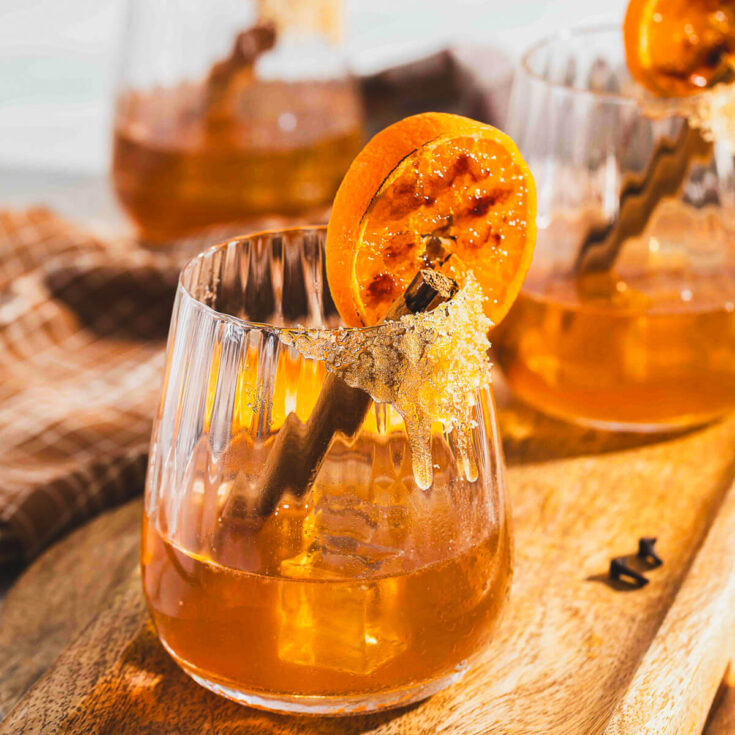 A brown bourbon old fashioned cocktails in a rocks glass garnished with an orange slice and a cinnamon stick.