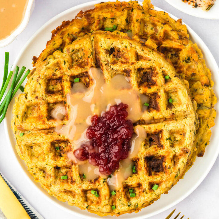 A white plate containing two crispy golden Stuffing Waffles topped with gravy and cranberry sauce.