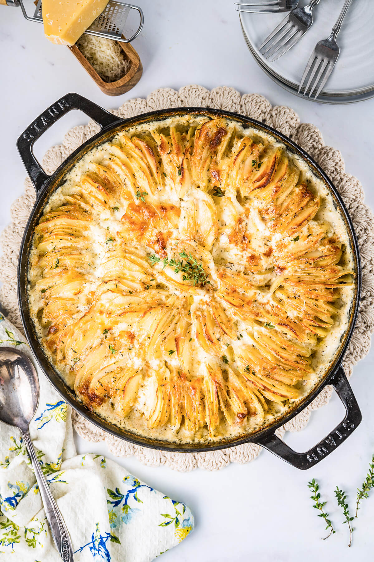 A large Staub baking dish containing golden baked cheesy potatoes au gratin and fresh thyme leaves.