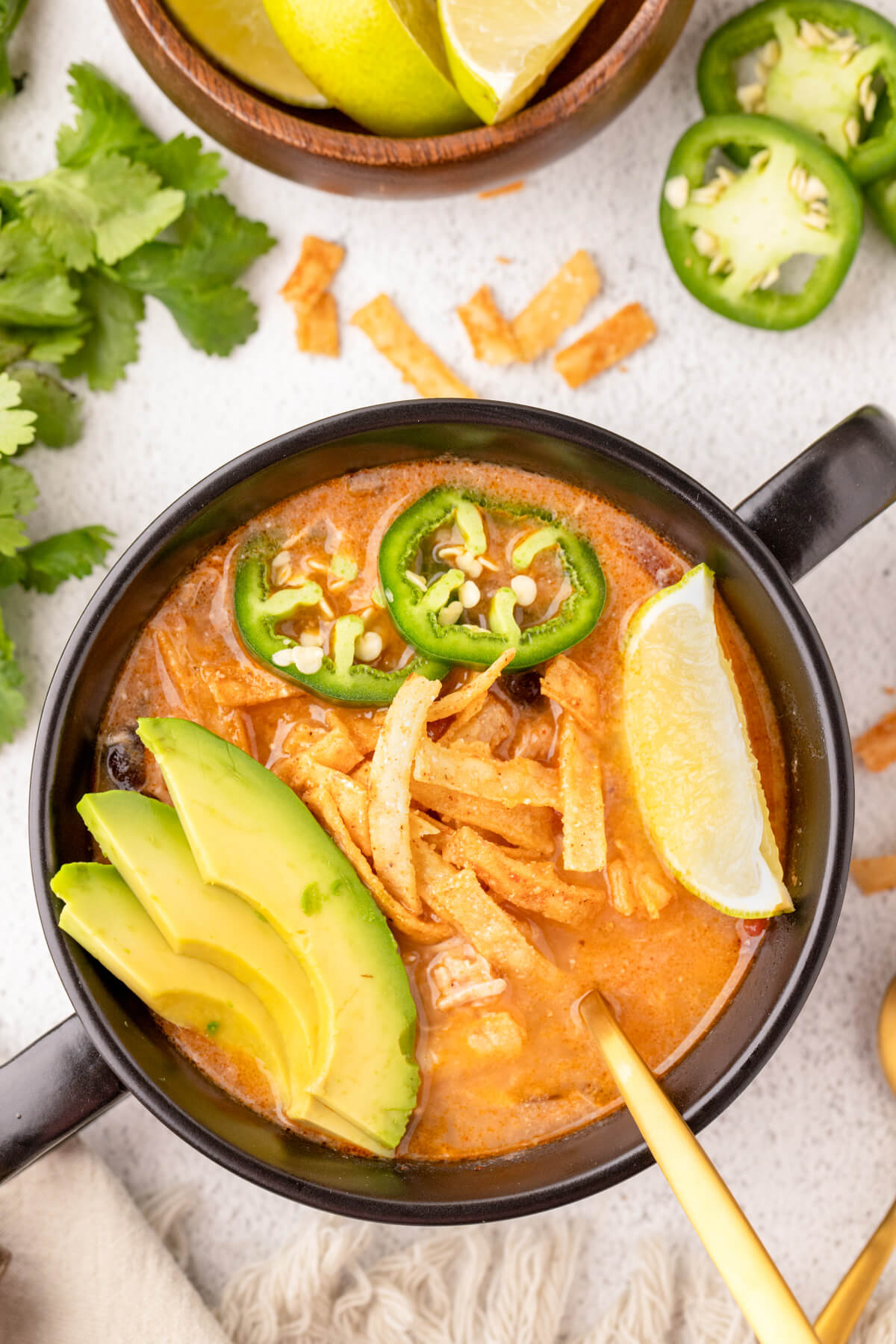 A black soup bowl full of creamy Chicken Enchilada Soup garnished with sliced avocados, tortilla strips, cilantro, lime, and jalapenos.