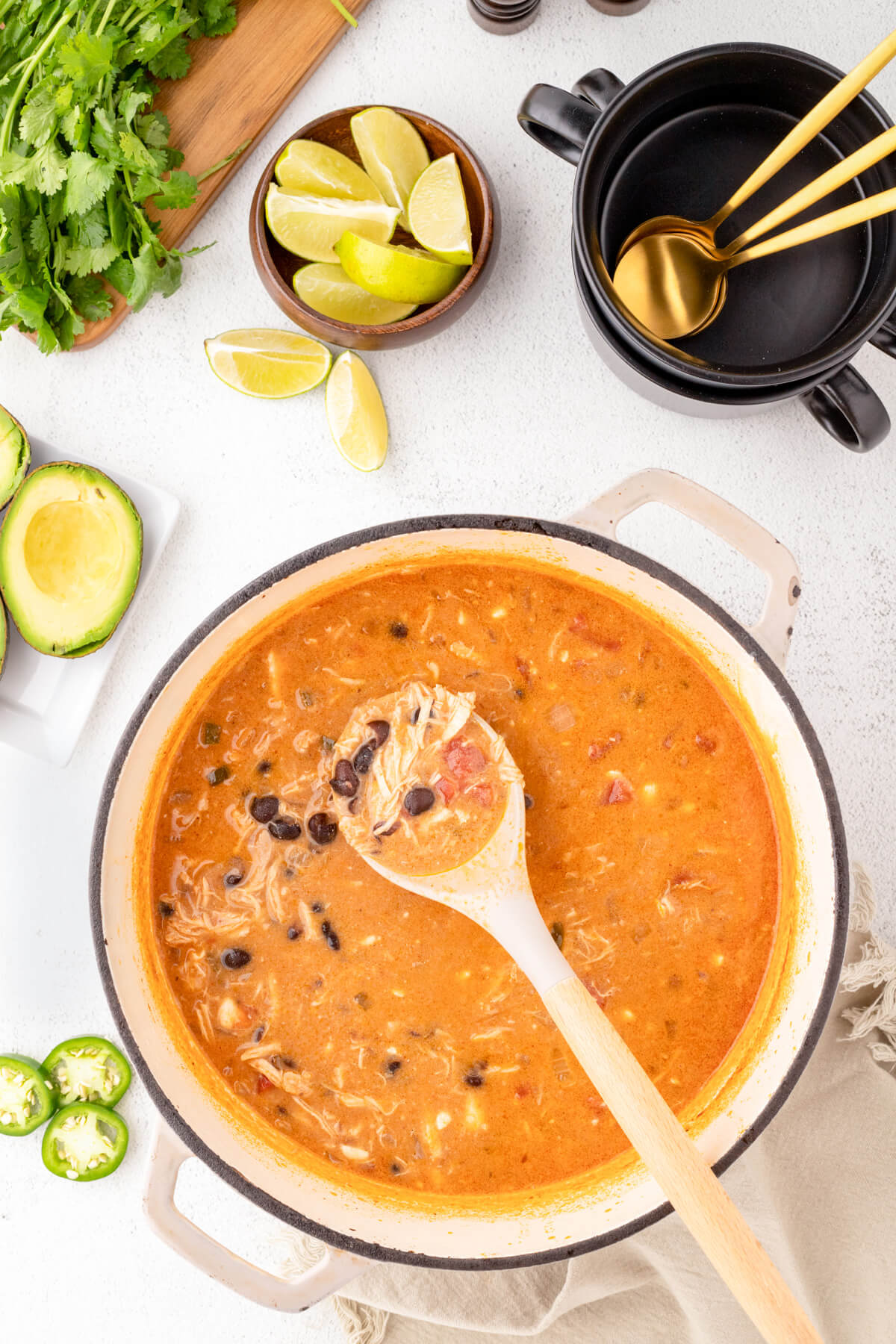 A soup pot full of Creamy Chicken Enchilada Soup surrounded by black soup bowls, spoons, and garnishes.