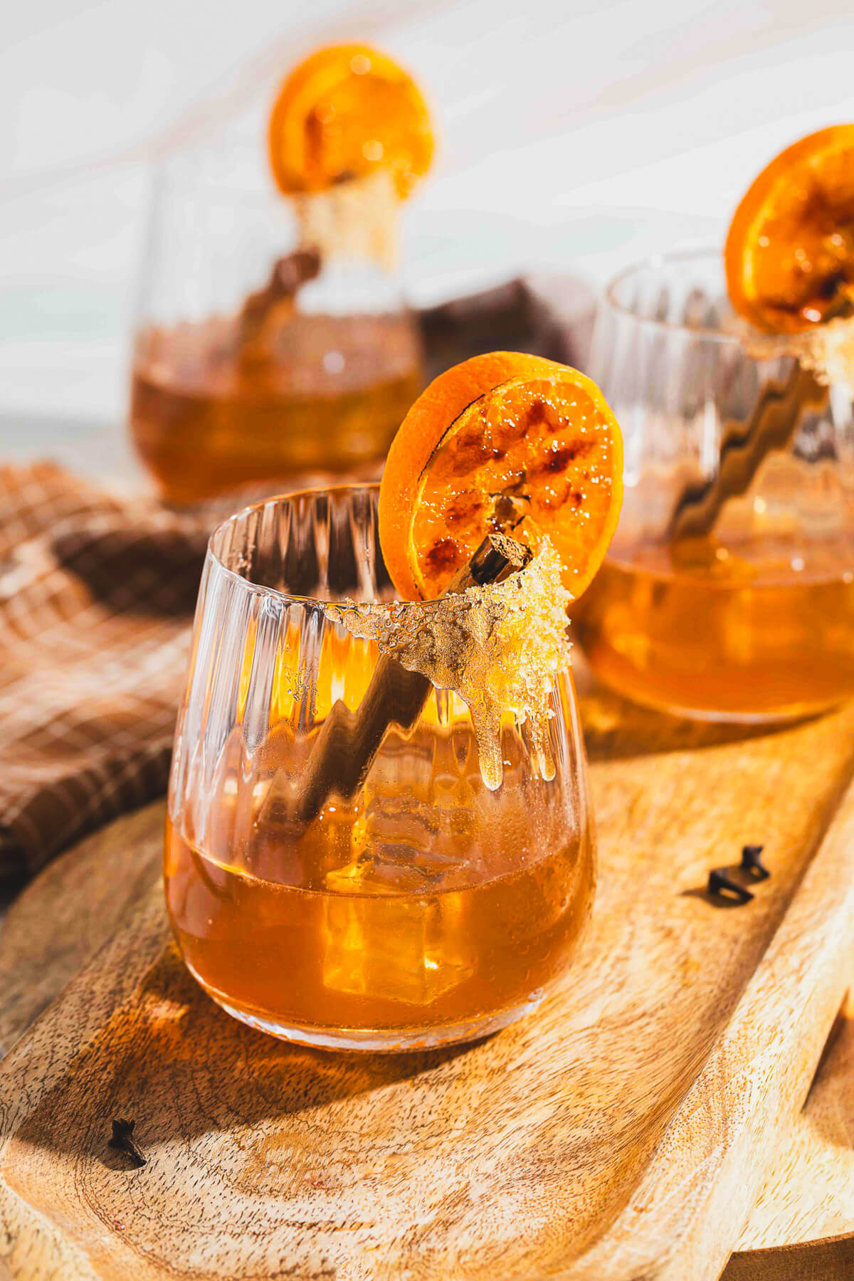 Three bourbon old fashioned cocktails in a rocks glasses garnished with orange slices and cinnamon sticks on a wooden board.