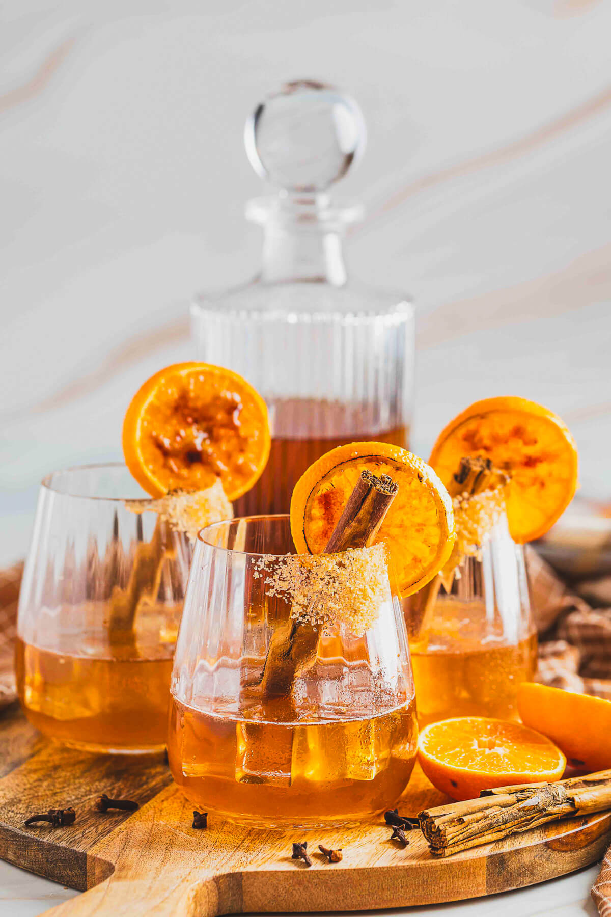 Three bourbon old fashioned cocktails in a rocks glasses in front of a decanter of bourbon on a wooden board.