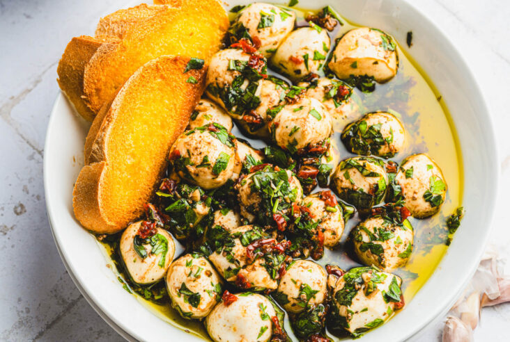 Two golden toasts in a white bowl filled with bite sized mozzarella balls coated in vibrant green herbs, yellow olive oil, and dotted with sundried tomatoes.